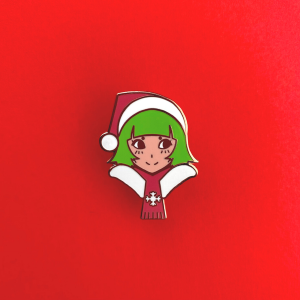 limited-edition holiday enamel pin by Ilya Kuvshinov. Christmas / Santa themed done in Ilya Kuvshinov's signature manga/anime-style. This manga / anime styled girl has green hair and wears a christmas hat and scarf. Only 200 pins were produced and is available at Plush Art Club.